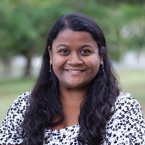 Smiling woman with long dark hair wearing a white floral blouse, outdoors in a park setting.