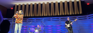 Female singer in a bright patterned shirt performs on stage with microphone in hand, drummer behind her and a bassist on the right under blue lighting.