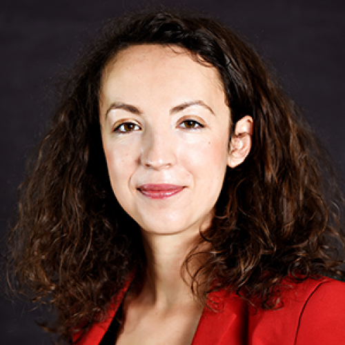 Portrait of a woman with shoulder-length curly brown hair wearing a red blazer against a dark studio background, with a subtle smile.