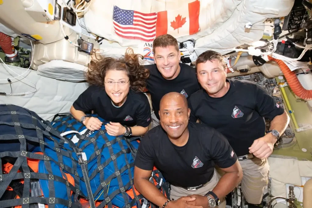 Four astronauts in black shirts pose for a crew photo inside a cramped space station module, with US and Canadian flags overhead and gear around them A blue-and-orange cargo bag visible in the foreground.